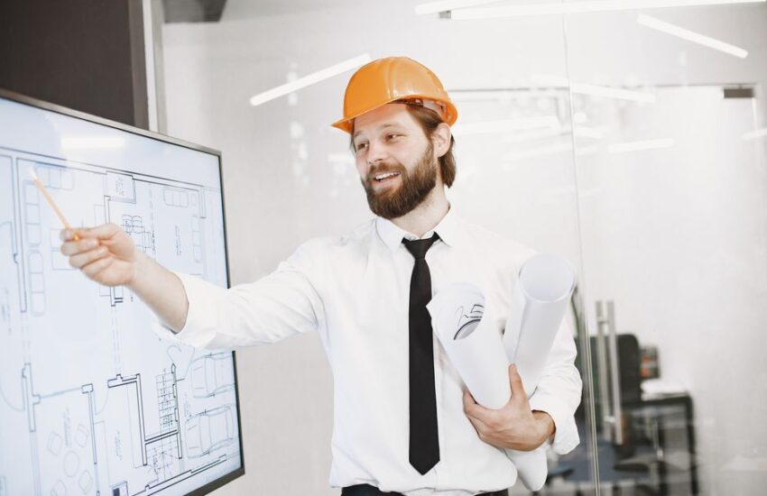 Engineer in helmet presenting architectural plans on a whiteboard in an office setting.