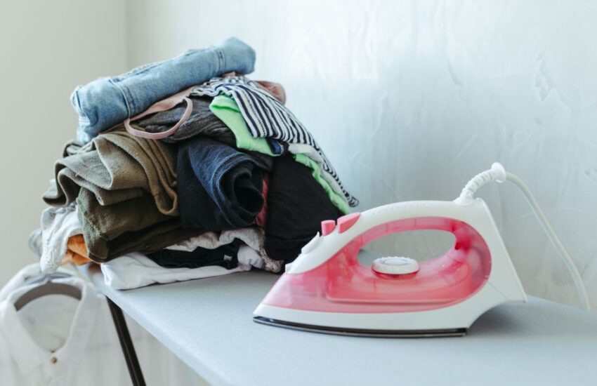 Tidy stack of clothes ready to be ironed with an iron on an ironing board.