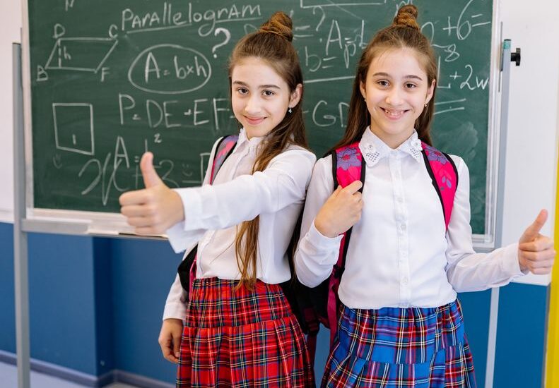 Two cheerful schoolgirls in uniforms giving thumbs up in front of a chalkboard filled with math equations.