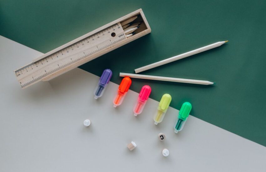 Flat lay of colorful highlighters and pencils with a wooden ruler on a green and white background.