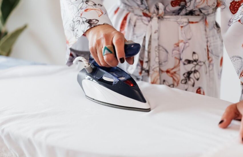 Crop unrecognizable female with manicured hands wearing classy silk robe and stylish ring ironing white sheets on ironing board