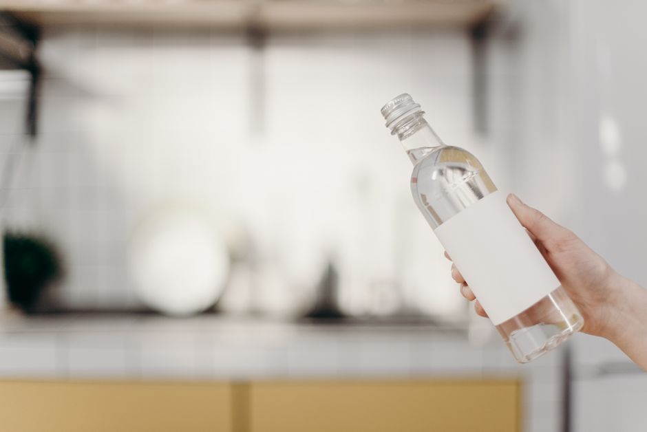 Hand holding a clear water bottle with blank label in a modern kitchen setting.