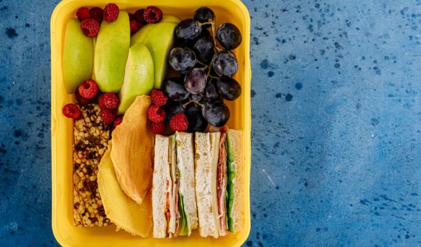 A vibrant lunchbox filled with sandwiches, apples, grapes, and raspberries on a blue background.