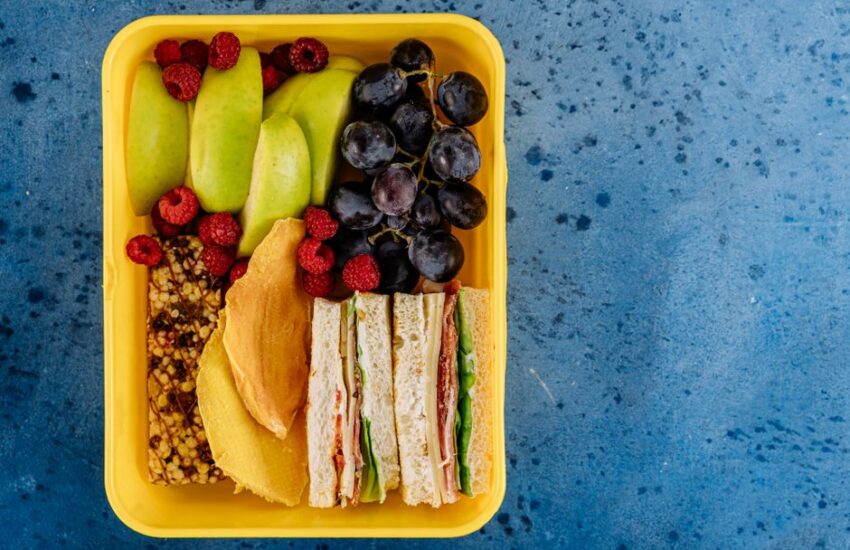 A vibrant lunchbox filled with sandwiches, apples, grapes, and raspberries on a blue background.
