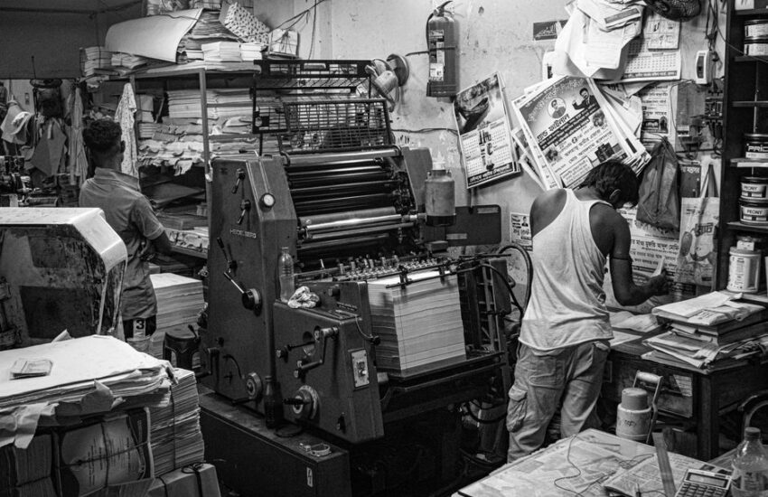 Black and white image of a bustling print shop with workers operating a printing press.
