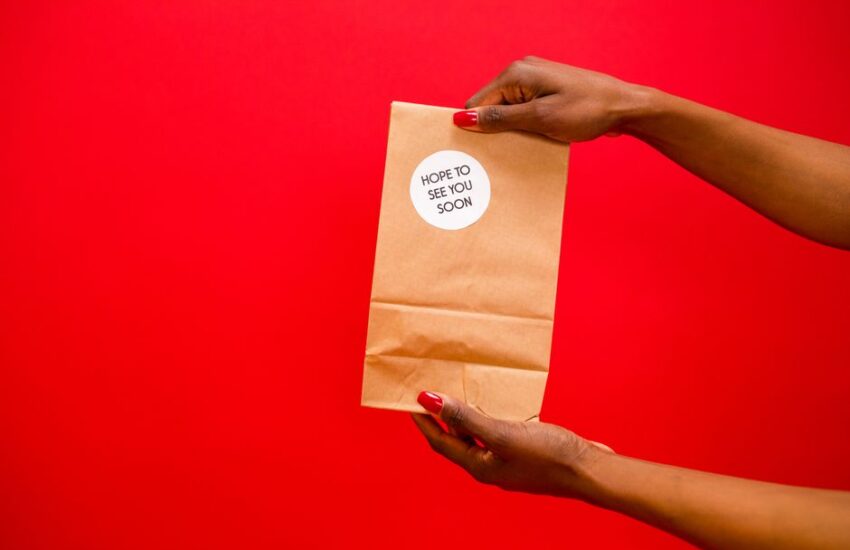 Two hands holding a paper bag with a 'Hope to see you soon' sticker, against a red background.
