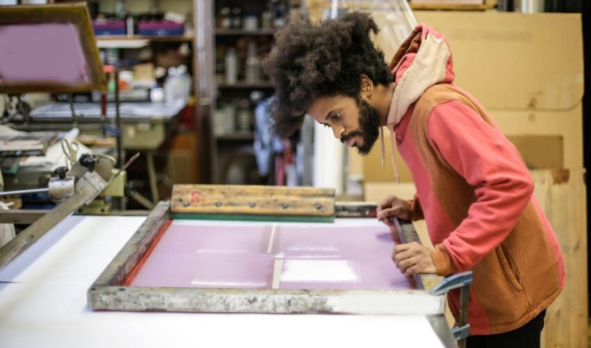 Side view of focused young bearded ethnic craftsman in casual hoodie standing near workbench with wooden frame and preparing colorful material in modern workshop