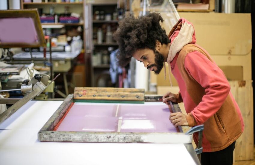 Side view of focused young bearded ethnic craftsman in casual hoodie standing near workbench with wooden frame and preparing colorful material in modern workshop