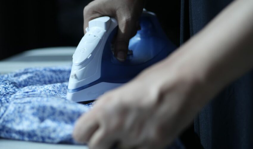 Detailed view of hands using an electric iron on blue fabric indoors.